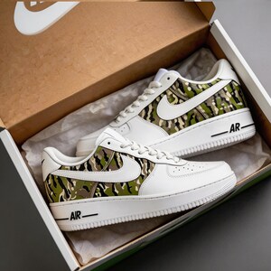 May include: White sneakers with a camouflage pattern on the side panels and heel. The shoes have a white sole and a large white Nike swoosh. The shoes are in a brown cardboard box with the Nike logo.