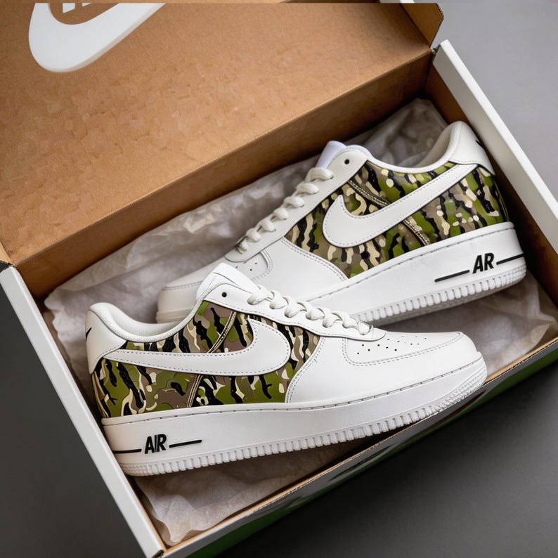 May include: White sneakers with a camouflage pattern on the side panels and heel. The shoes have a white sole and a large white Nike swoosh. The shoes are in a brown cardboard box with the Nike logo.