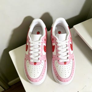 May include: Pair of white and pink sneakers with a strawberry theme. The shoes feature a white base with pink accents and red strawberry designs. The sneakers have white laces and soles, and are decorated with small strawberry graphics.