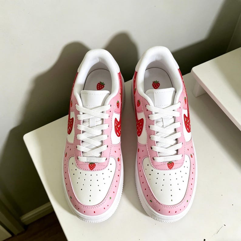 May include: Pair of white and pink sneakers with a strawberry theme. The shoes feature a white base with pink accents and red strawberry designs. The sneakers have white laces and soles, and are decorated with small strawberry graphics.