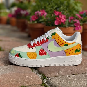 Puede incluir: Zapatillas Air Force 1 blancas personalizadas con un dise&ntilde;o de frutas colorido. La zapatilla presenta fresas, pi&ntilde;as y corazones en rojo, amarillo y verde, con un logo Nike verde claro. La suela es blanca, sobre una superficie de ladrillo.