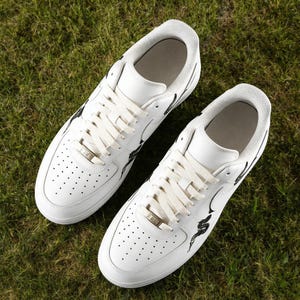 May include: A pair of white sneakers with black graphic accents. The shoes have white laces, a perforated toe box, and a silver metal lace tag. The sneakers are set on a bed of green grass.