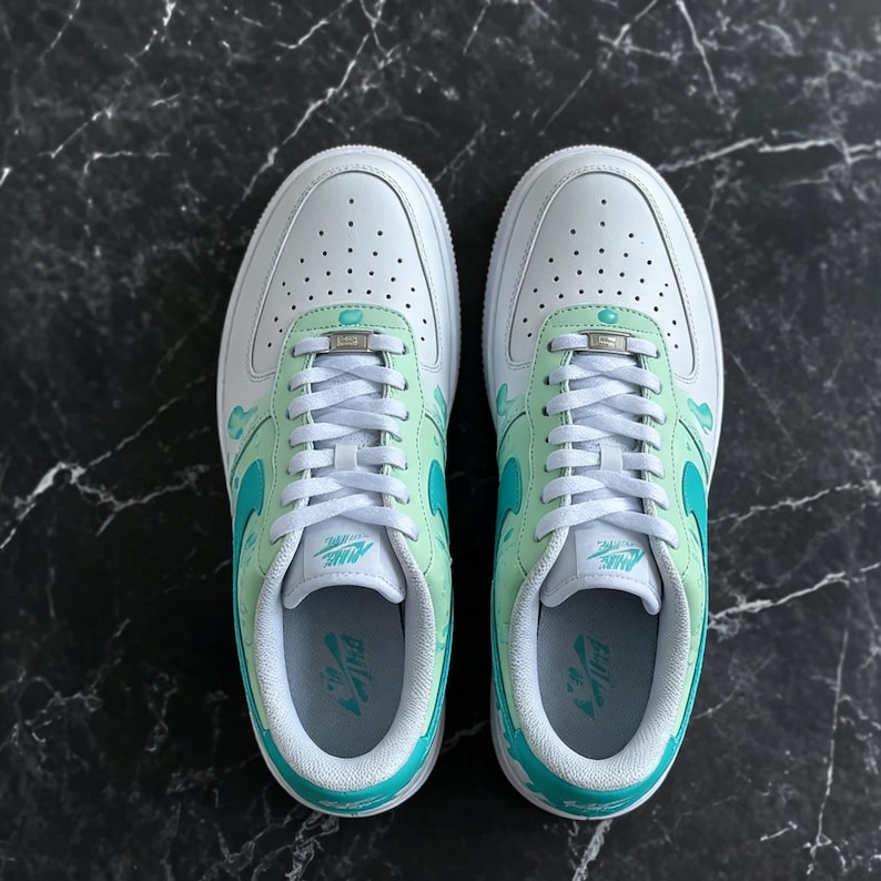 May include: Pair of white sneakers with mint green and teal accents. The shoes feature a classic design with a perforated toe box, teal swoosh, and mint green detailing on the tongue and heel. White laces and soles complete the look.