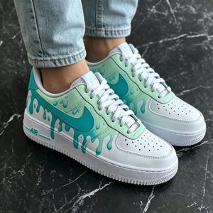 May include: White and teal sneakers with a unique drip design. The shoes feature a white base with teal accents, including the iconic swoosh and a dripping paint effect. The word "AIR" is visible on the side.