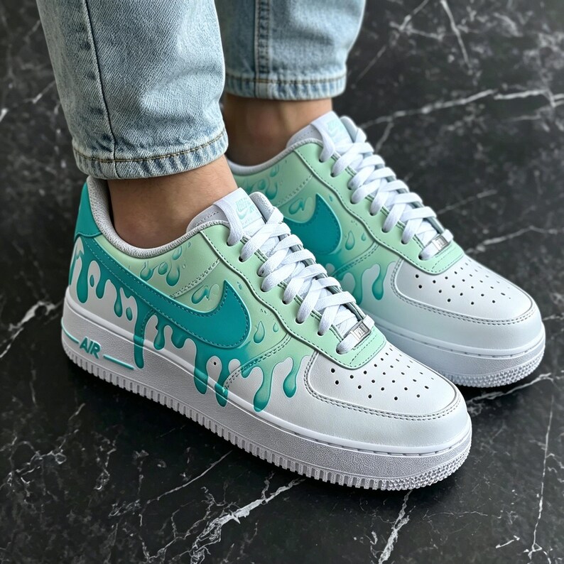May include: White and teal sneakers with a unique drip design. The shoes feature a white base with teal accents, including the iconic swoosh and a dripping paint effect. The word "AIR" is visible on the side.