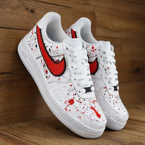 Może przedstawiać: Białe trampki z czerwonymi i czarnymi rozpryskami farby. Buty mają czerwone logo swoosh z boku i białe sznurowadła. Podeszwy są białe, a trampki są prezentowane na drewnianej powierzchni.