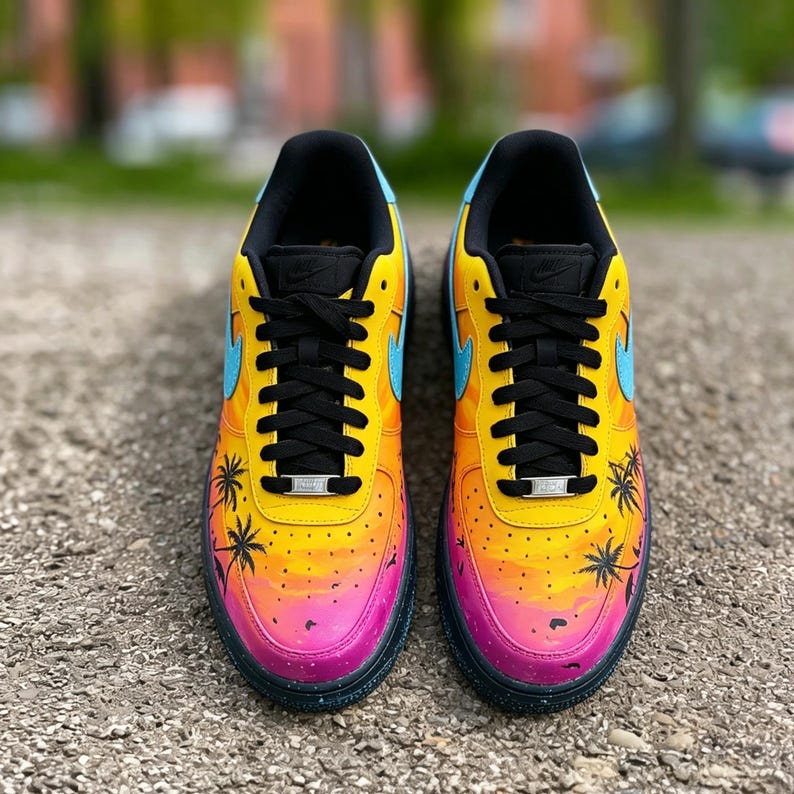 May include: Pair of custom-painted sneakers featuring a sunset design. The shoes have a yellow, orange, and pink gradient with black palm tree silhouettes. They have black laces, a black sole, and a blue Nike swoosh.