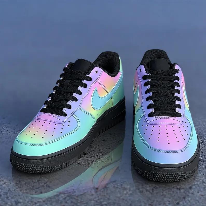 May include: Pair of iridescent sneakers with a gradient of colors, including pink, blue, and yellow. The shoes have black laces, a black sole, and a Nike swoosh logo on the side. The sneakers are displayed on a reflective surface.