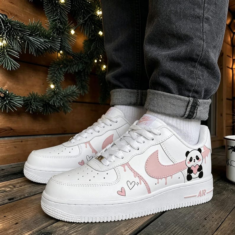 Puede incluir: Zapatillas blancas con detalles en rosa, incluyendo un logotipo Swoosh, efecto de pintura goteando y detalles de corazones. Un gr&aacute;fico de panda sosteniendo un coraz&oacute;n est&aacute; en el lateral. Los zapatos tienen suela y cordones blancos.