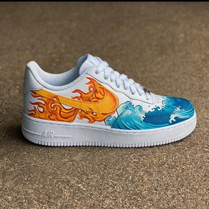 May include: White athletic shoe with custom artwork. The design features a yellow and orange flame on the side, and a blue and white wave pattern. The shoe has white laces and a white sole. The word "AIR" is visible on the side of the sole.
