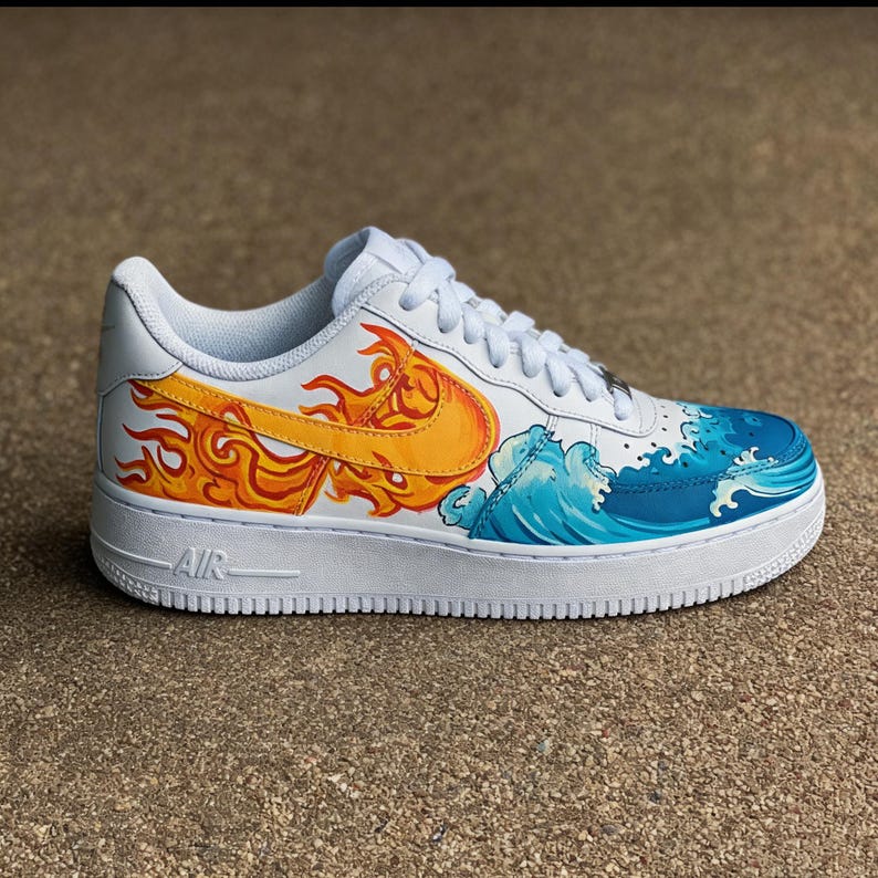 May include: White athletic shoe with custom artwork. The design features a yellow and orange flame on the side, and a blue and white wave pattern. The shoe has white laces and a white sole. The word "AIR" is visible on the side of the sole.