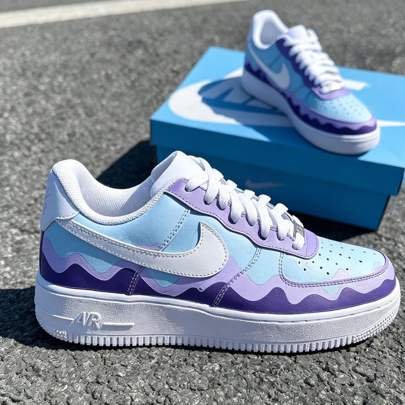 May include: Customized Air Force 1 sneakers in shades of blue and purple with a wavy design. The shoes have white laces, a white sole, and a white Nike swoosh. A matching pair sits on a blue box.