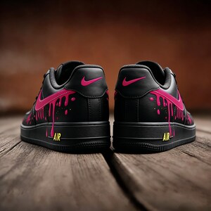 May include: Pair of black sneakers with pink dripping paint design. The shoes feature a pink Nike swoosh and the word "AIR" in yellow. The sneakers are set against a wooden background.