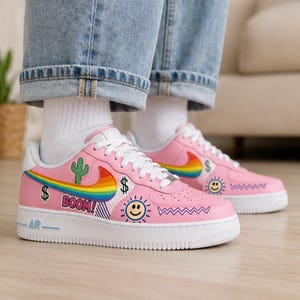 May include: Pink sneakers with white soles and laces. The shoes feature a rainbow swoosh, a cactus, dollar signs, a smiling sun, and the word "BOOM!" in a speech bubble. The shoes are worn with blue jeans and white socks.