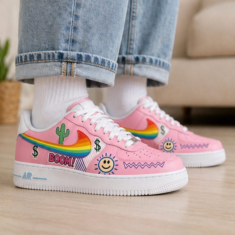 May include: Pink sneakers with white soles and laces. The shoes feature a rainbow swoosh, a cactus, dollar signs, a smiling sun, and the word "BOOM!" in a speech bubble. The shoes are worn with blue jeans and white socks.