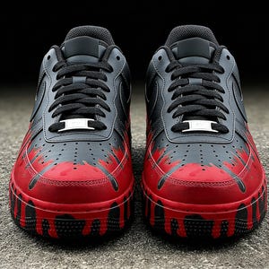 May include: Pair of black and red sneakers with a unique design. The shoes feature a black upper with red accents that appear to drip down the sides. Black laces and a white metal tag complete the look. The shoes are on a gray surface.