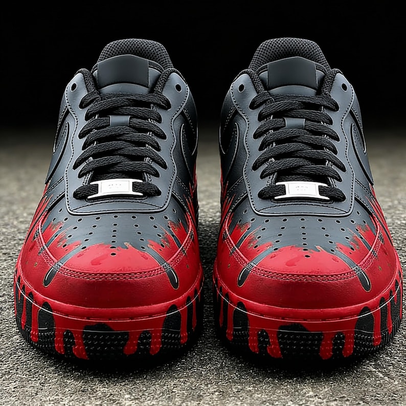 May include: Pair of black and red sneakers with a unique design. The shoes feature a black upper with red accents that appear to drip down the sides. Black laces and a white metal tag complete the look. The shoes are on a gray surface.
