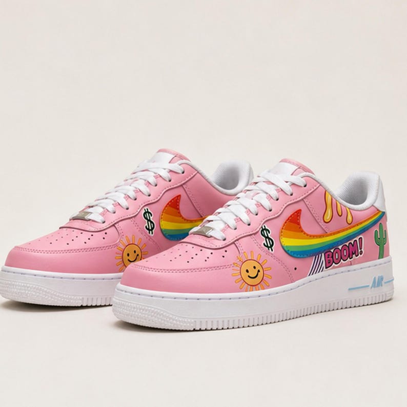 May include: Pink sneakers with white soles and laces. The shoes feature a rainbow swoosh, dollar signs, smiling suns, and the word "BOOM!" with a cactus graphic. A stylish and colorful design.