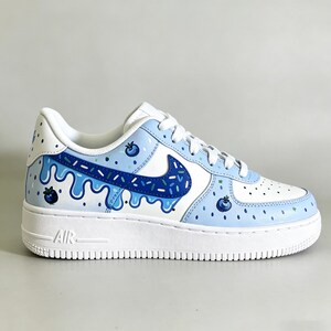 May include: White and light blue Air Force 1 sneakers with a blueberry-themed design. The shoe features a white sole, a blue Nike swoosh with a sprinkle pattern, and blueberry illustrations.