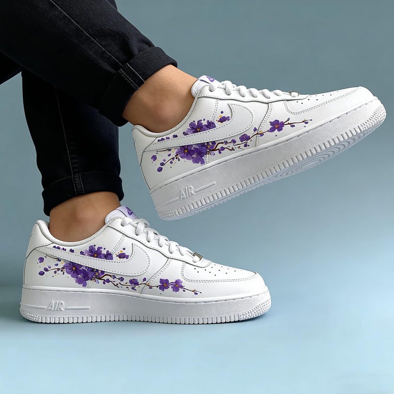 May include: White sneakers with purple floral design. The shoes feature a classic low-top silhouette with a white sole and the word "AIR" on the side. The purple floral pattern adds a unique touch to the footwear.
