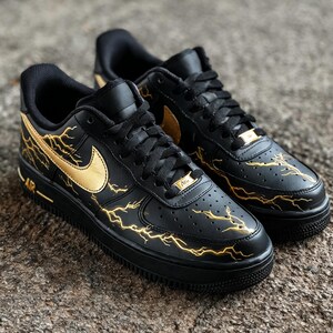 Op de afbeelding: Paar zwarte Nike Air Force 1 sneakers met gouden bliksemontwerpen en gouden Nike swoosh logo's. De schoenen hebben zwarte veters en zolen, met een gouden Nike tag op de veters.