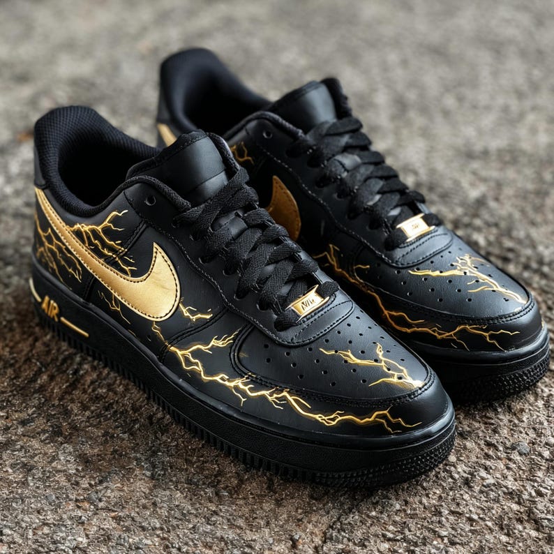 Op de afbeelding: Paar zwarte Nike Air Force 1 sneakers met gouden bliksemontwerpen en gouden Nike swoosh logo's. De schoenen hebben zwarte veters en zolen, met een gouden Nike tag op de veters.
