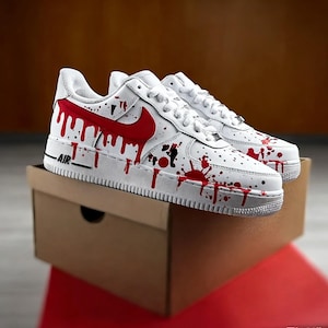 Op de afbeelding: Witte sneakers met een rood druipverf ontwerp. De schoenen hebben een rode Nike swoosh en zwarte spetters. Het woord "AIR" staat in het zwart op de zijkant van de witte zool. De sneakers worden gepresenteerd op een bruine kartonnen doos.