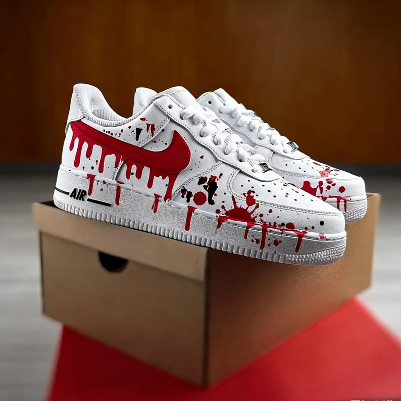 Op de afbeelding: Witte sneakers met een rood druipverf ontwerp. De schoenen hebben een rode Nike swoosh en zwarte spetters. Het woord "AIR" staat in het zwart op de zijkant van de witte zool. De sneakers worden gepresenteerd op een bruine kartonnen doos.