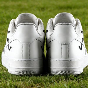 May include: A pair of white sneakers with black decorative accents, viewed from the back. The shoes are set against a backdrop of green grass, highlighting their clean design and contrasting details. The soles have a textured pattern.