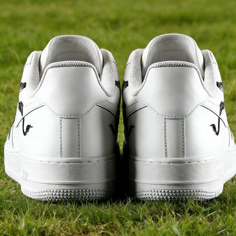 May include: A pair of white sneakers with black decorative accents, viewed from the back. The shoes are set against a backdrop of green grass, highlighting their clean design and contrasting details. The soles have a textured pattern.