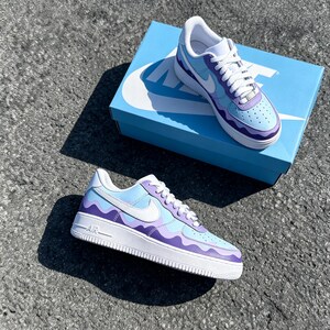 May include: A pair of custom-painted sneakers with a wavy design in shades of light blue and purple. The shoes have white laces, a white sole, and a white Nike swoosh. The shoes are displayed on a light blue Nike box.