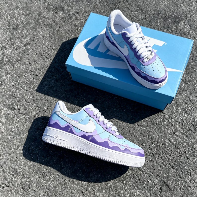 May include: A pair of custom-painted sneakers with a wavy design in shades of light blue and purple. The shoes have white laces, a white sole, and a white Nike swoosh. The shoes are displayed on a light blue Nike box.