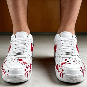 Op de afbeelding: Witte sneakers met rode accenten en een spetterverf ontwerp. De schoenen hebben een rood swoosh-logo en rode verfspetters op de neus en zijkanten. Witte veters en witte zolen.