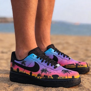 May include: Customized black sneakers with a gradient of blue, purple, and pink. The shoes feature a black Nike swoosh and a design of palm trees and a cityscape. The soles are black.
