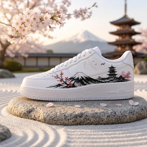 May include: White sneaker with a Japanese-inspired design featuring Mount Fuji, a pagoda, and cherry blossoms. The shoe has a white sole and the Nike swoosh logo. The design is in black and red.