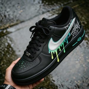 May include: Black Nike Air Force 1 sneaker with custom spiderweb and dripping paint design. The shoe features a silver swoosh, green and yellow paint drips, and a white spiderweb graphic on the side. The word "AIR" is in teal.