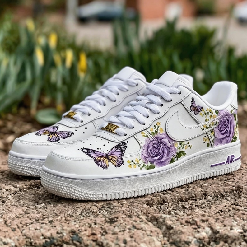 Op de afbeelding: Witte sneakers met vlinder- en bloemmotieven. De schoenen hebben paarse rozen, gele accenten en vlindergraphics. De zolen zijn wit met een paarse streep en het woord "AR".