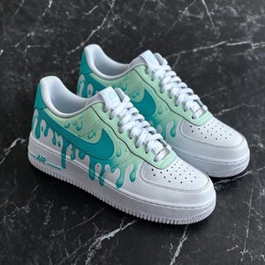 May include: White and teal Air Force 1 sneakers with a drip design. The shoes feature a white base with teal accents, including the Nike swoosh and a dripping paint effect. The laces are white.