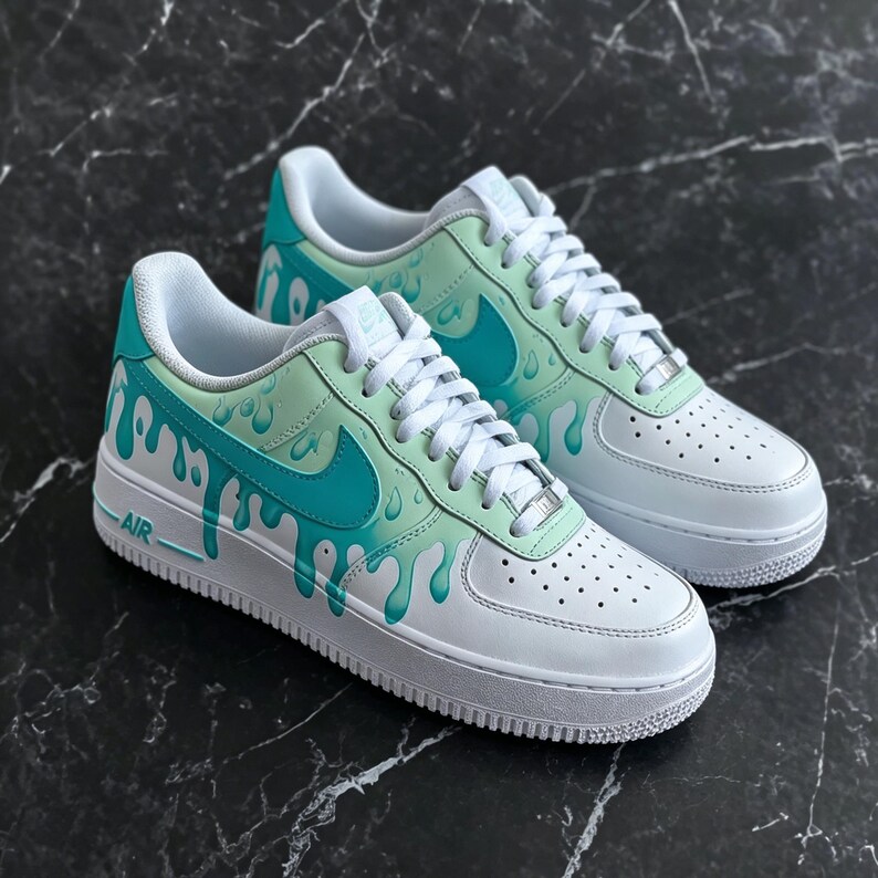 May include: White and teal Air Force 1 sneakers with a drip design. The shoes feature a white base with teal accents, including the Nike swoosh and a dripping paint effect. The laces are white.