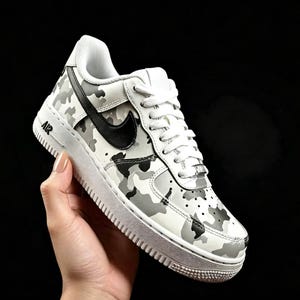 May include: White and gray camouflage pattern sneaker with a black swoosh logo. The shoe has white laces and a white sole. The word "AIR" is printed in black on the side of the shoe.