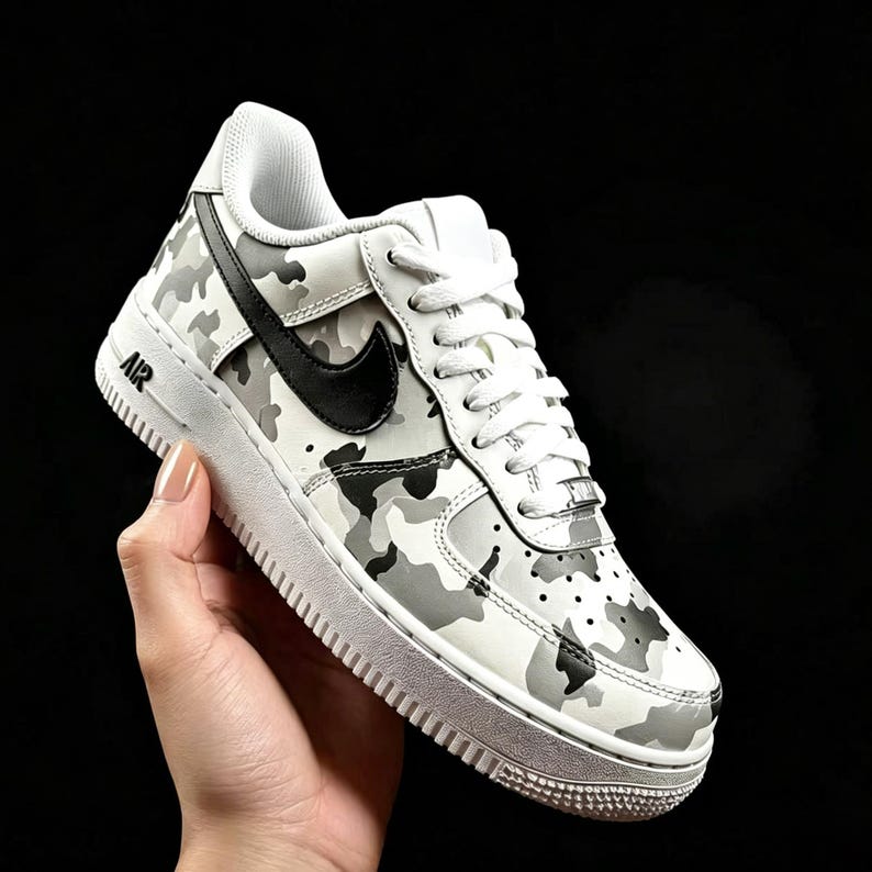 May include: White and gray camouflage pattern sneaker with a black swoosh logo. The shoe has white laces and a white sole. The word "AIR" is printed in black on the side of the shoe.