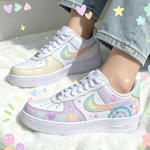 May include: White sneakers with pastel rainbow and heart designs. The shoes feature a white sole and laces, with a colorful swoosh logo. One shoe has a yellow and pastel design, while the other has a rainbow and moon design.