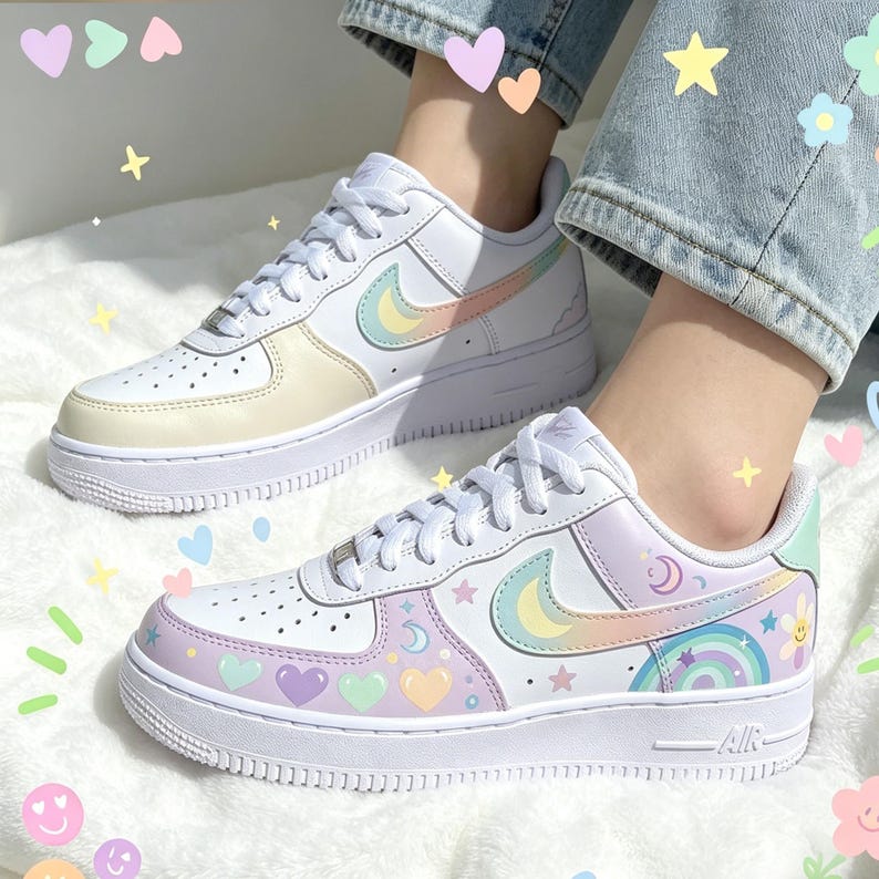 May include: White sneakers with pastel rainbow and heart designs. The shoes feature a white sole and laces, with a colorful swoosh logo. One shoe has a yellow and pastel design, while the other has a rainbow and moon design.