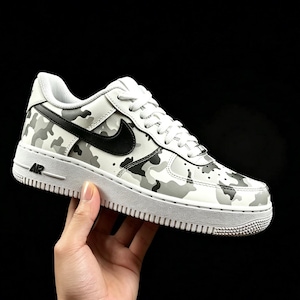 May include: White sneakers with a camouflage pattern in shades of gray and black. The shoes feature a black swoosh logo and white laces. The sole is white with the word "AIR" visible.