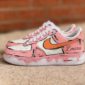May include: Customized pink and white sneakers with an orange swoosh and black outline detailing. The shoes feature a dripping paint effect and the name "Lmara" on the side. White laces and a white sole complete the design.