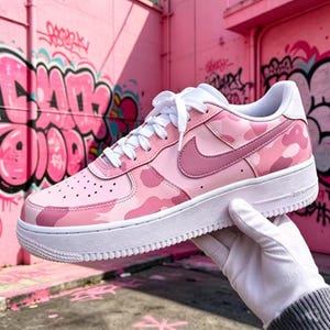 May include: Pink camouflage sneakers with a white sole and laces. The Nike swoosh is a darker shade of pink. The shoe is held by a gloved hand in front of a pink wall with graffiti.