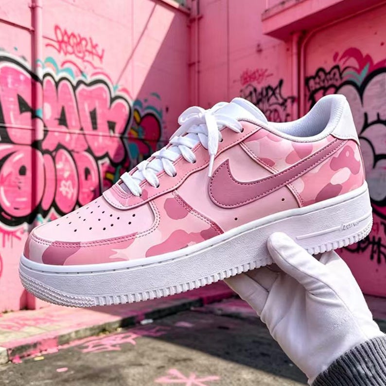 May include: Pink camouflage sneakers with a white sole and laces. The Nike swoosh is a darker shade of pink. The shoe is held by a gloved hand in front of a pink wall with graffiti.