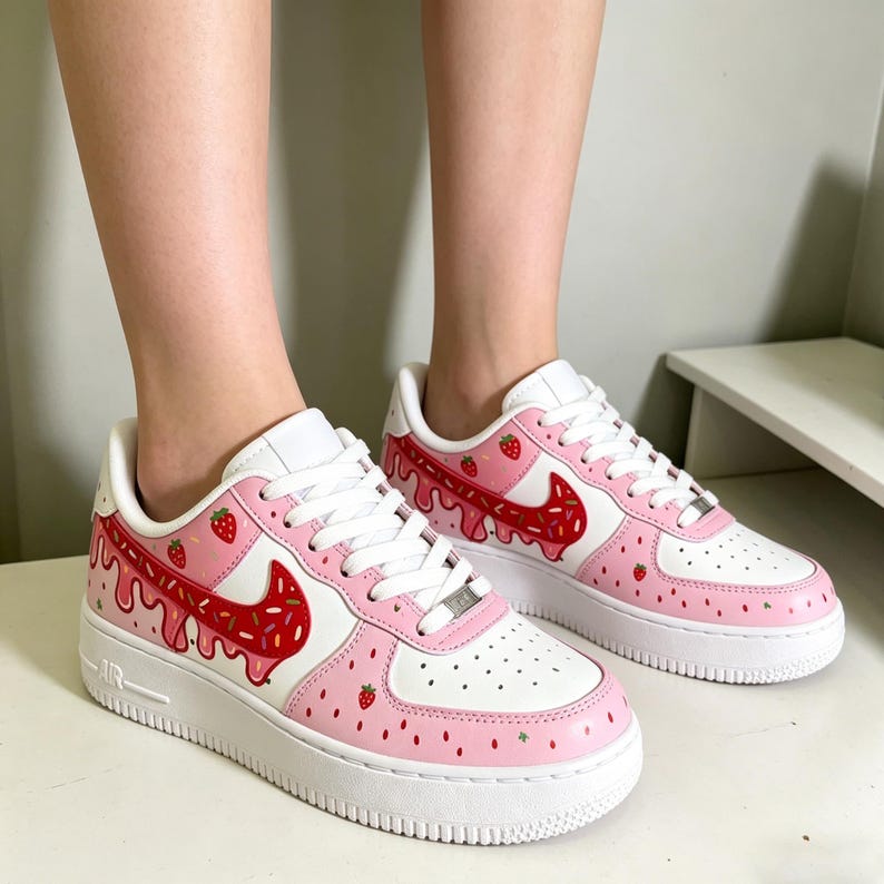 May include: White and pink sneakers with a strawberry theme. The shoes feature a white base with pink accents, a red swoosh, and strawberry details. The soles are white.