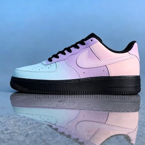 Op de afbeelding: Een enkele Air Force 1 sneaker in een pastelkleurverloop van lichtblauw, lavendel en roze. De schoen heeft een zwarte zool, veters en hielstuk. De iconische Nike swoosh is zichtbaar aan de zijkant.