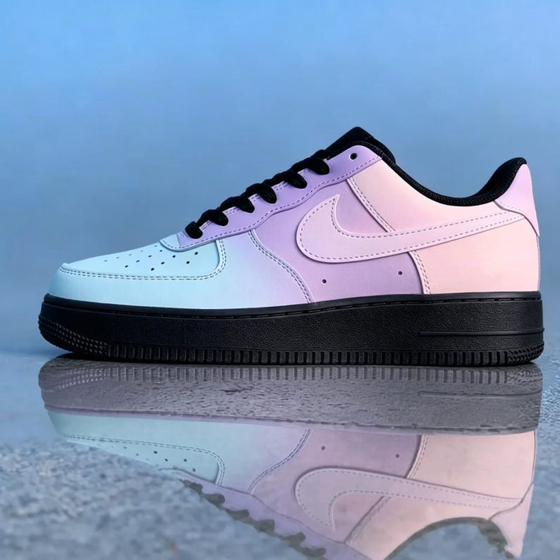Op de afbeelding: Een enkele Air Force 1 sneaker in een pastelkleurverloop van lichtblauw, lavendel en roze. De schoen heeft een zwarte zool, veters en hielstuk. De iconische Nike swoosh is zichtbaar aan de zijkant.